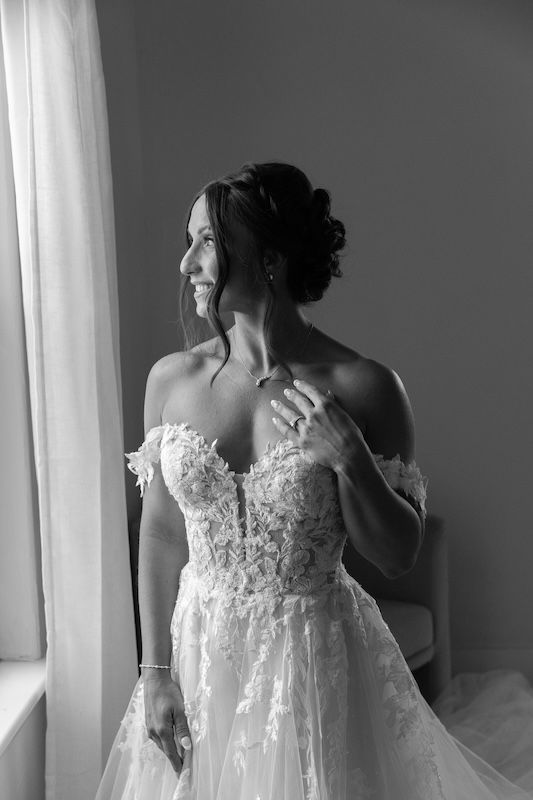 Black and white photo of a smiling bride in an off-shoulder lace wedding gown standing by a window.