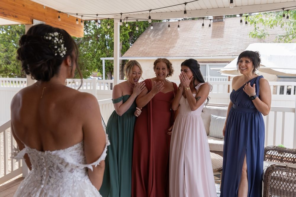 Bridesmaids in colorful dresses react with joy and tears seeing the bride in her wedding gown for the first time.