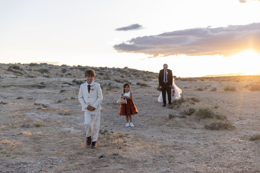 Groom and flower girl walk through desert landscape at sunset during outdoor wedding ceremony.
