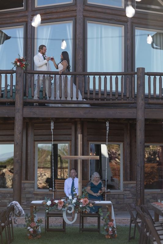Couple on rustic log cabin balcony during wedding ceremony with guests seated below near floral cross.