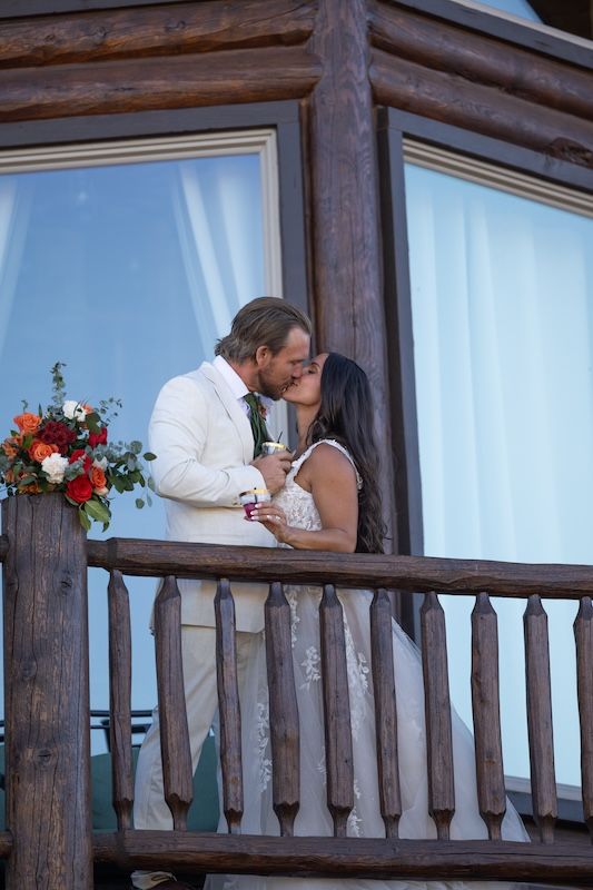 Bride and groom share a romantic kiss on a rustic wooden balcony, with a red floral bouquet nearby.