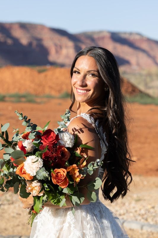 Smiling bride in lace dress holds vibrant red and orange bouquet with red rock desert landscape in background.