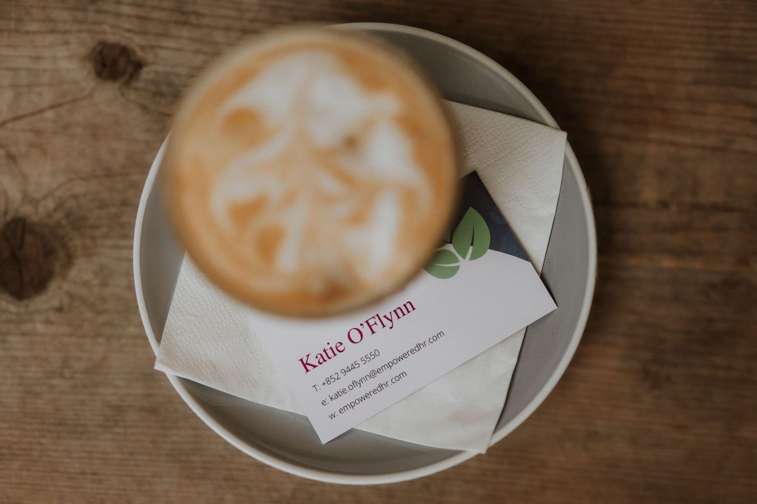 A latte with latte art sits on a ceramic plate beside a Katie O'Flynn business card on a wooden table.