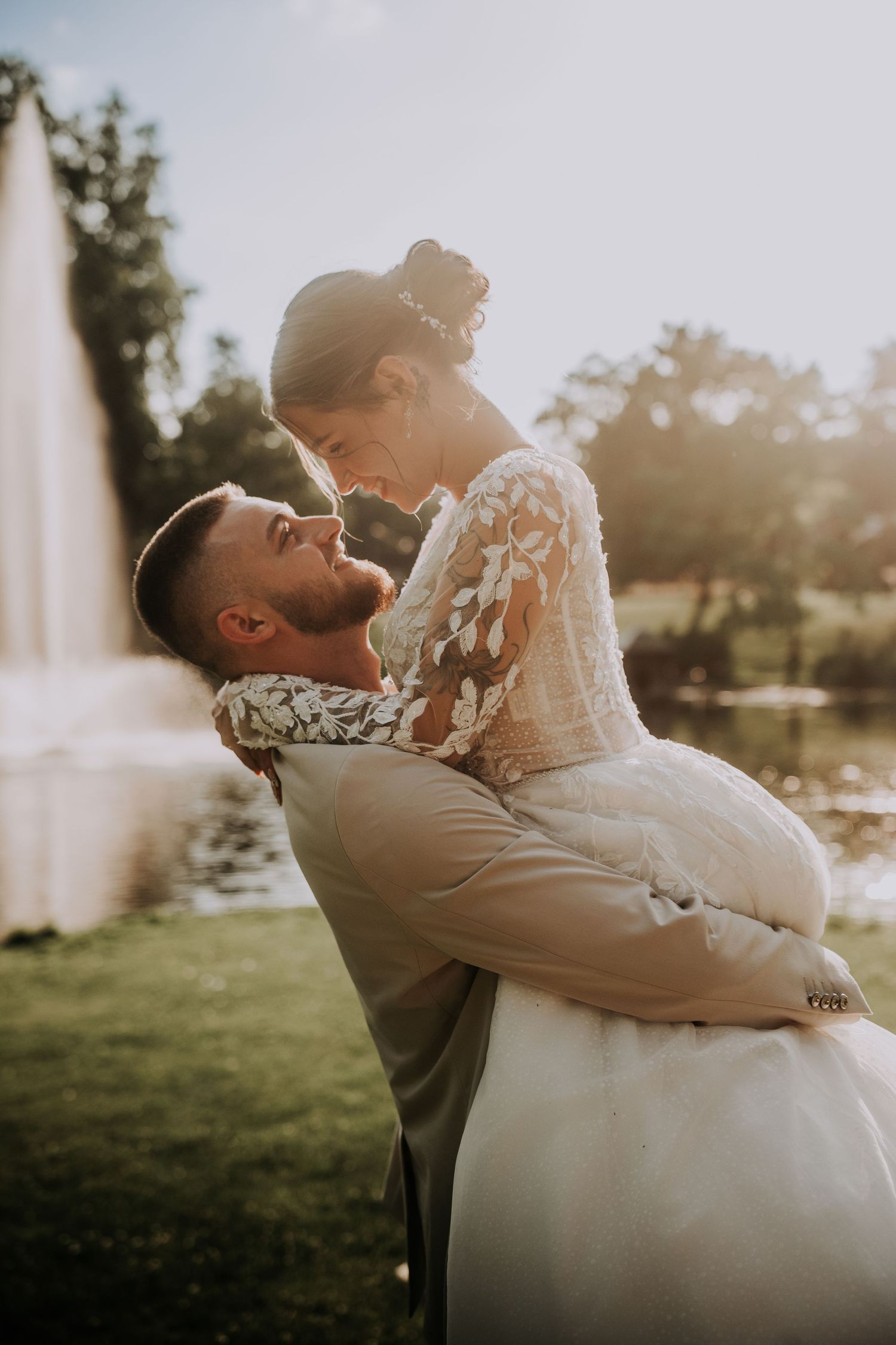 Groom lifting bride in wedding attire near a sunlit fountain outdoors.