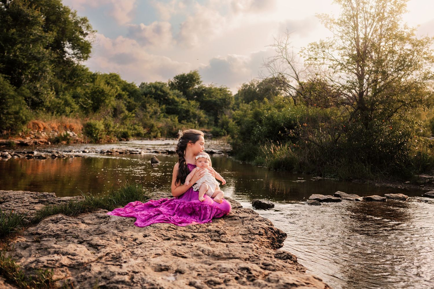 A girl in a flowing purple dress sits on riverside rocks, tenderly cradling a baby in soft natural light.