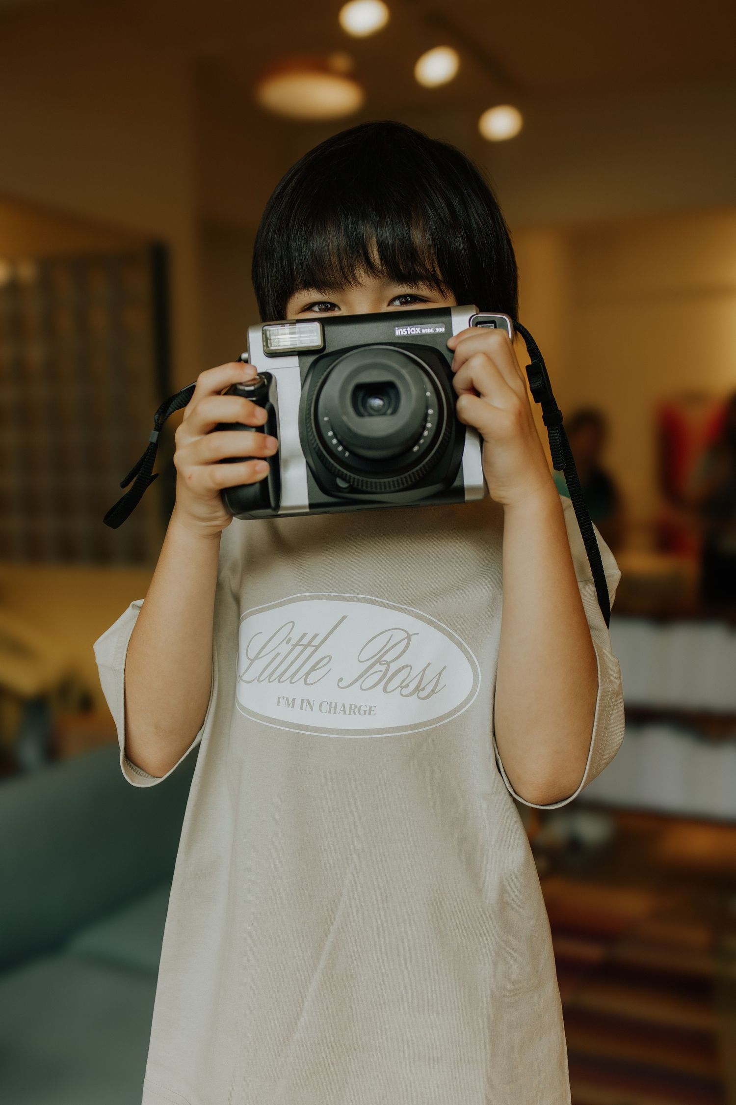 A young child wearing a Little Boss shirt holds a Fujifilm Instax wide camera up to their face indoors.