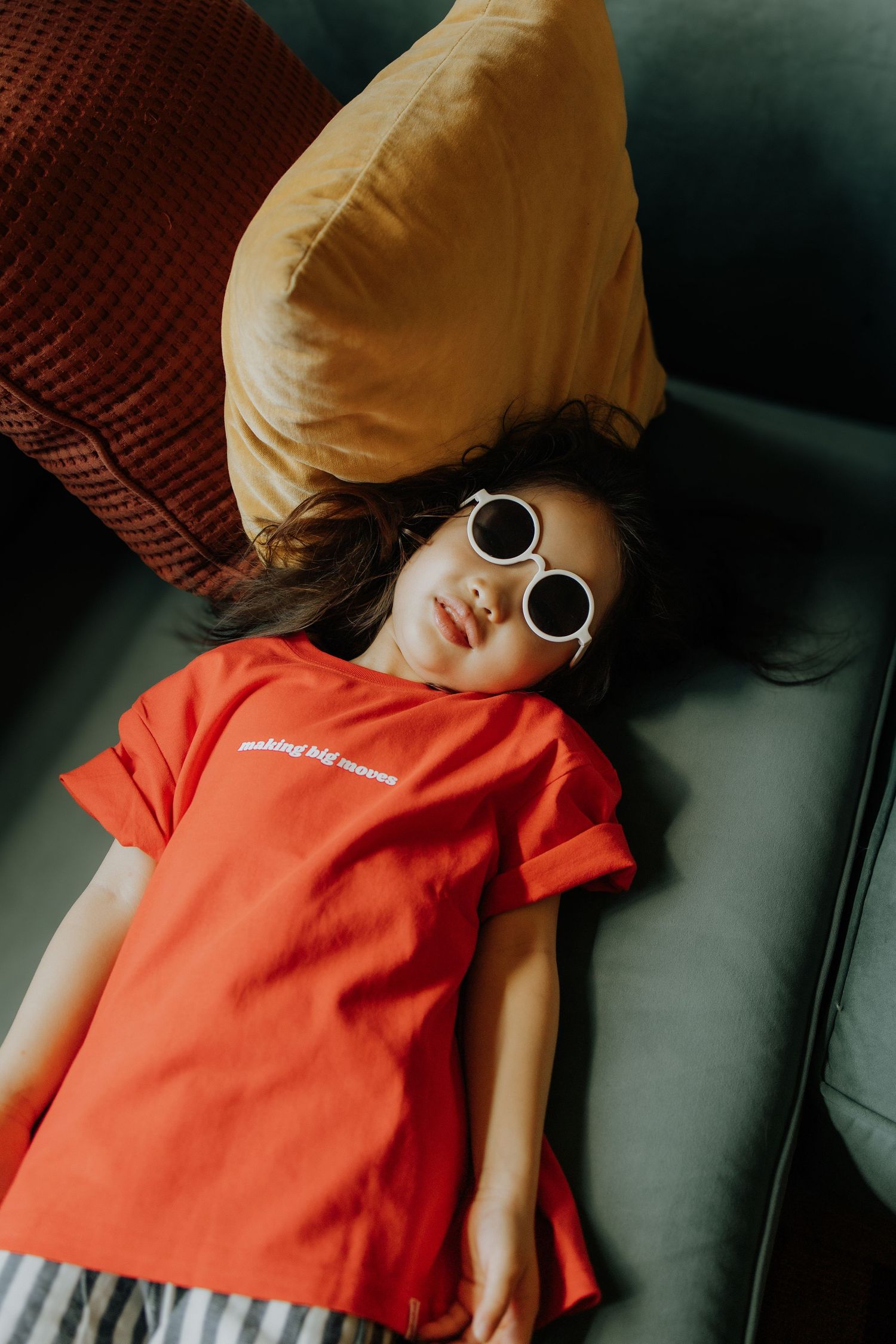 Child in red t-shirt and white sunglasses lying on a couch with colorful pillows.