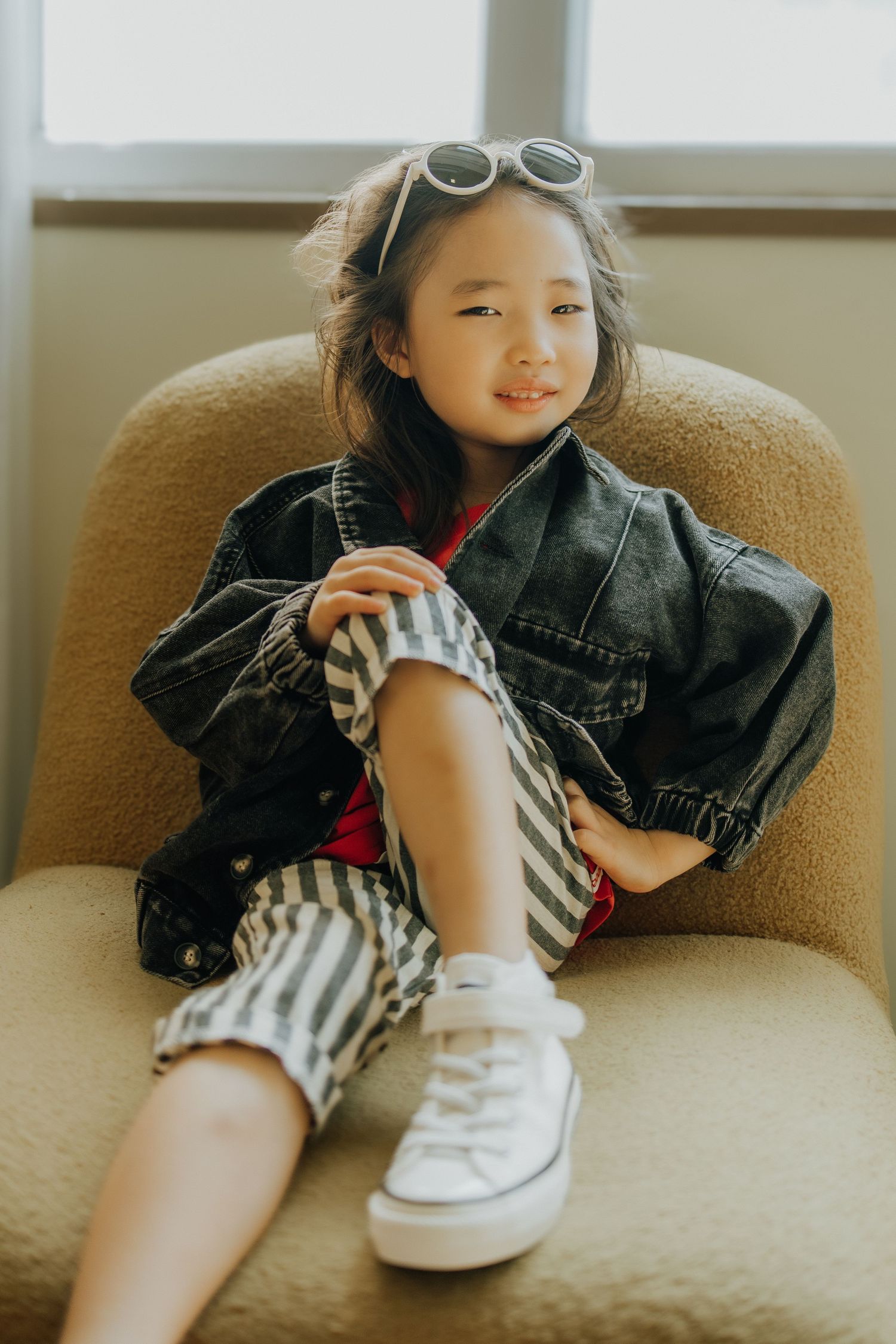Young girl in denim jacket, striped shorts, and sunglasses sitting relaxed in a beige armchair smiling.