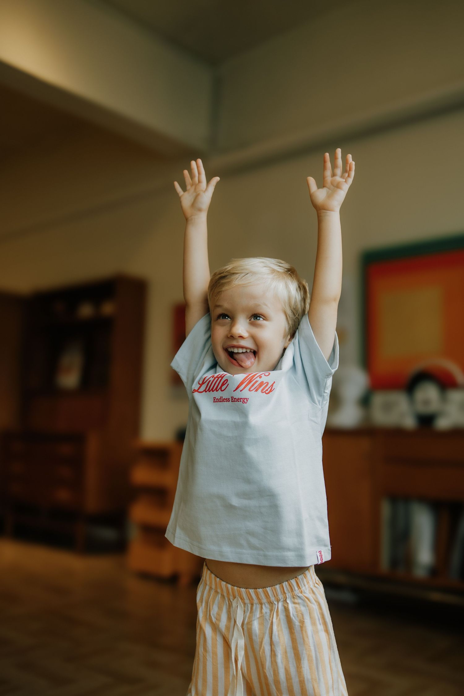 Excited young child in a light blue graphic tee raises both arms up with a big smile indoors.