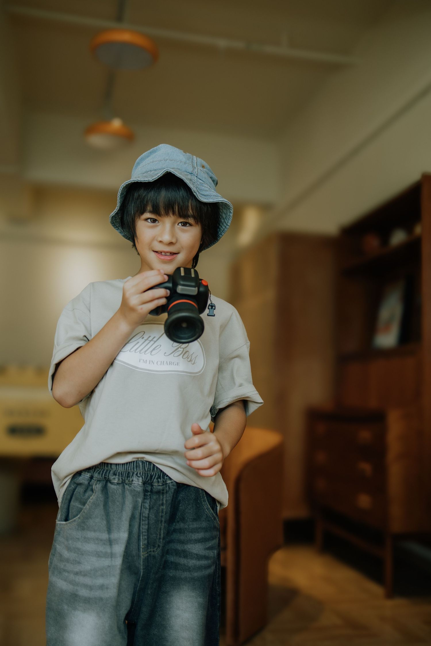 A young boy wearing a bucket hat holds a camera indoors, smiling confidently in a casual white tee and jeans.