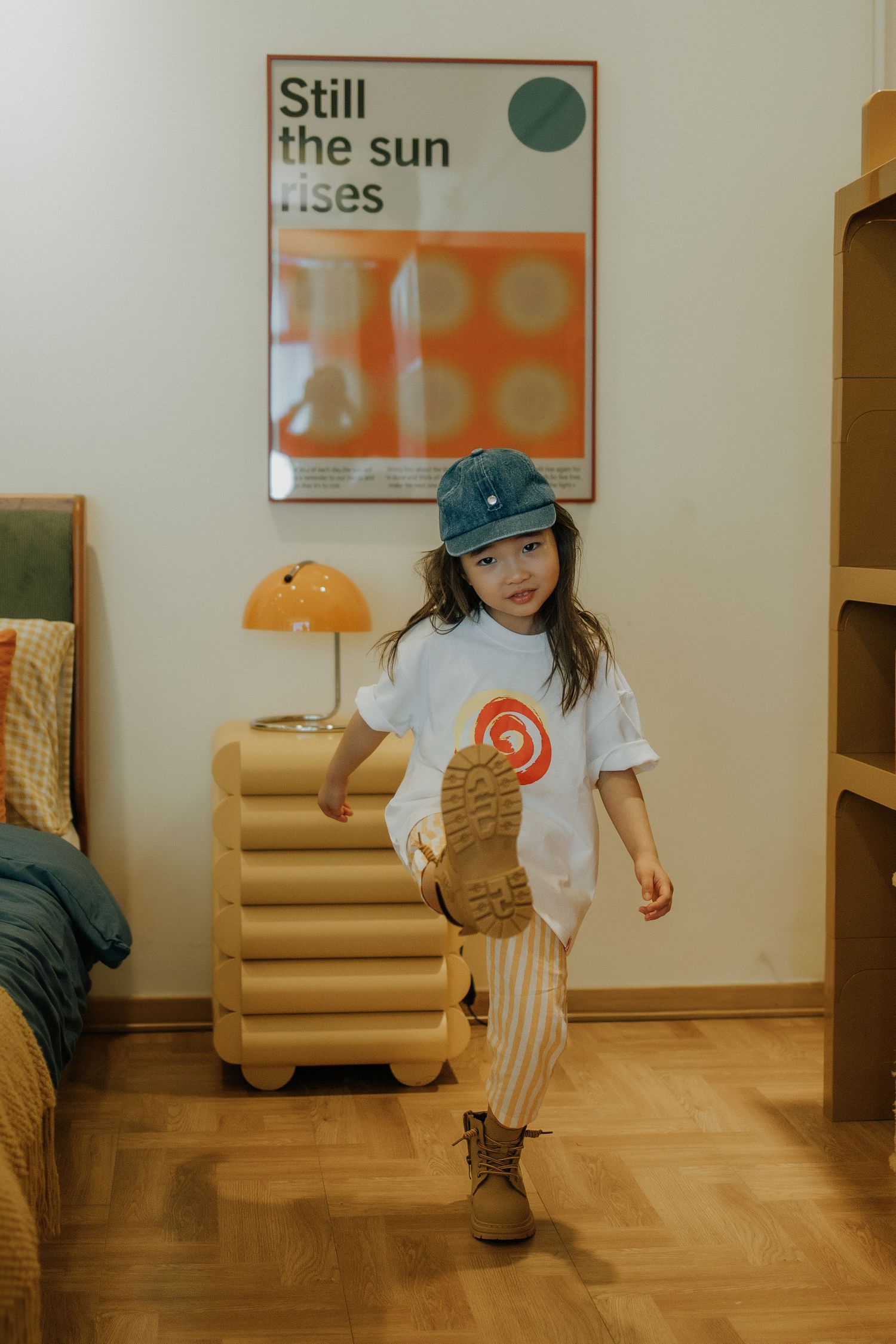 Young girl in white tee, striped pants, and blue cap poses playfully in a cozy room with retro decor.