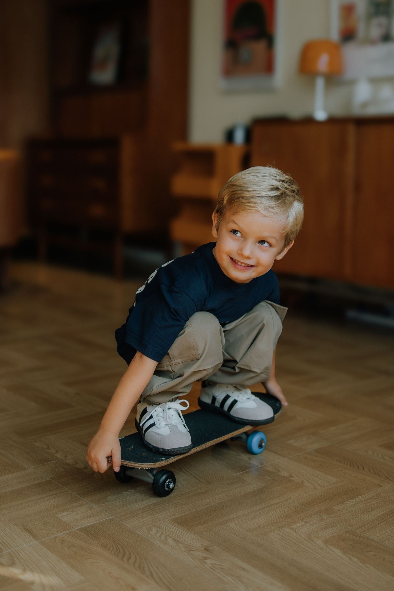 Young blonde boy smiling while crouching on a skateboard indoors on a wooden herringbone floor.