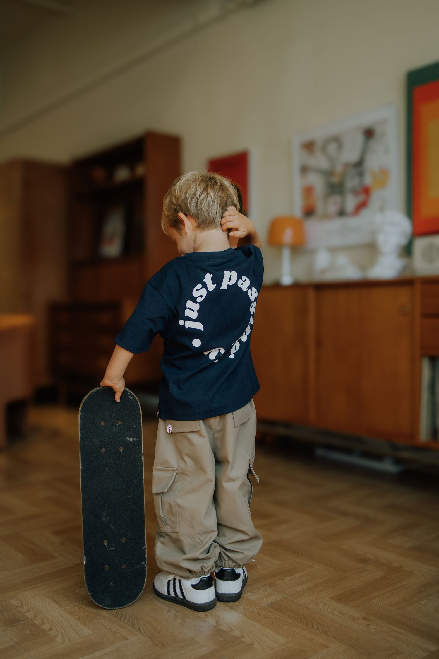 Young boy holding a skateboard indoors, wearing a navy graphic tee and khaki cargo pants with Adidas sneakers.