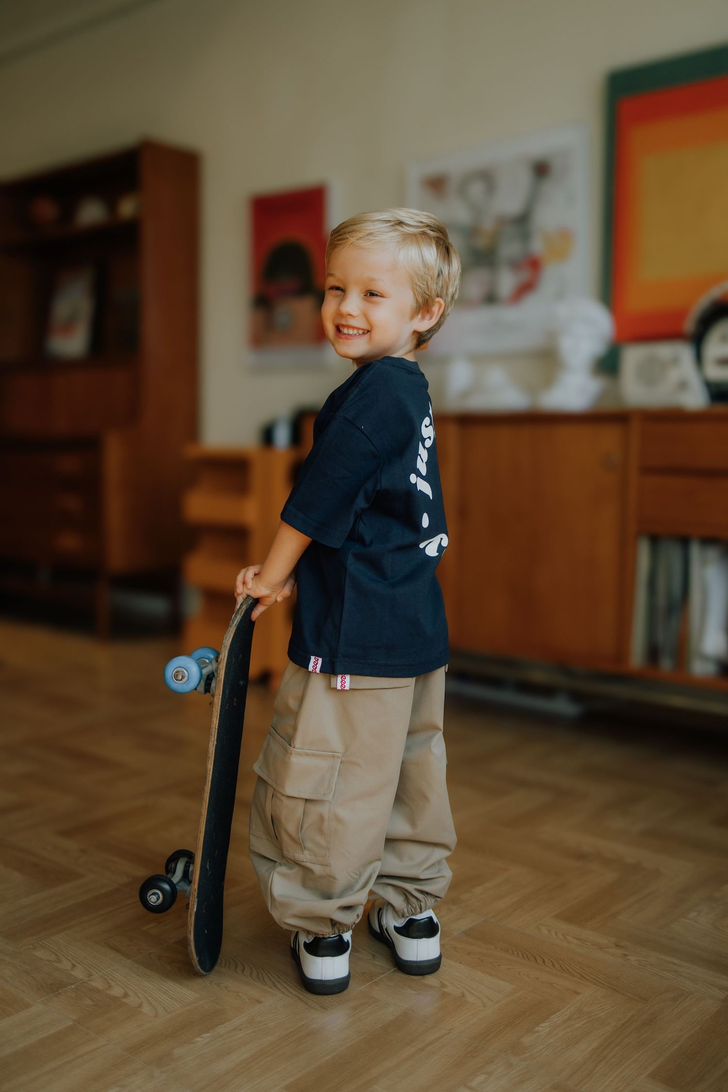 Smiling toddler boy holding a skateboard indoors, wearing a navy t-shirt and khaki cargo pants.