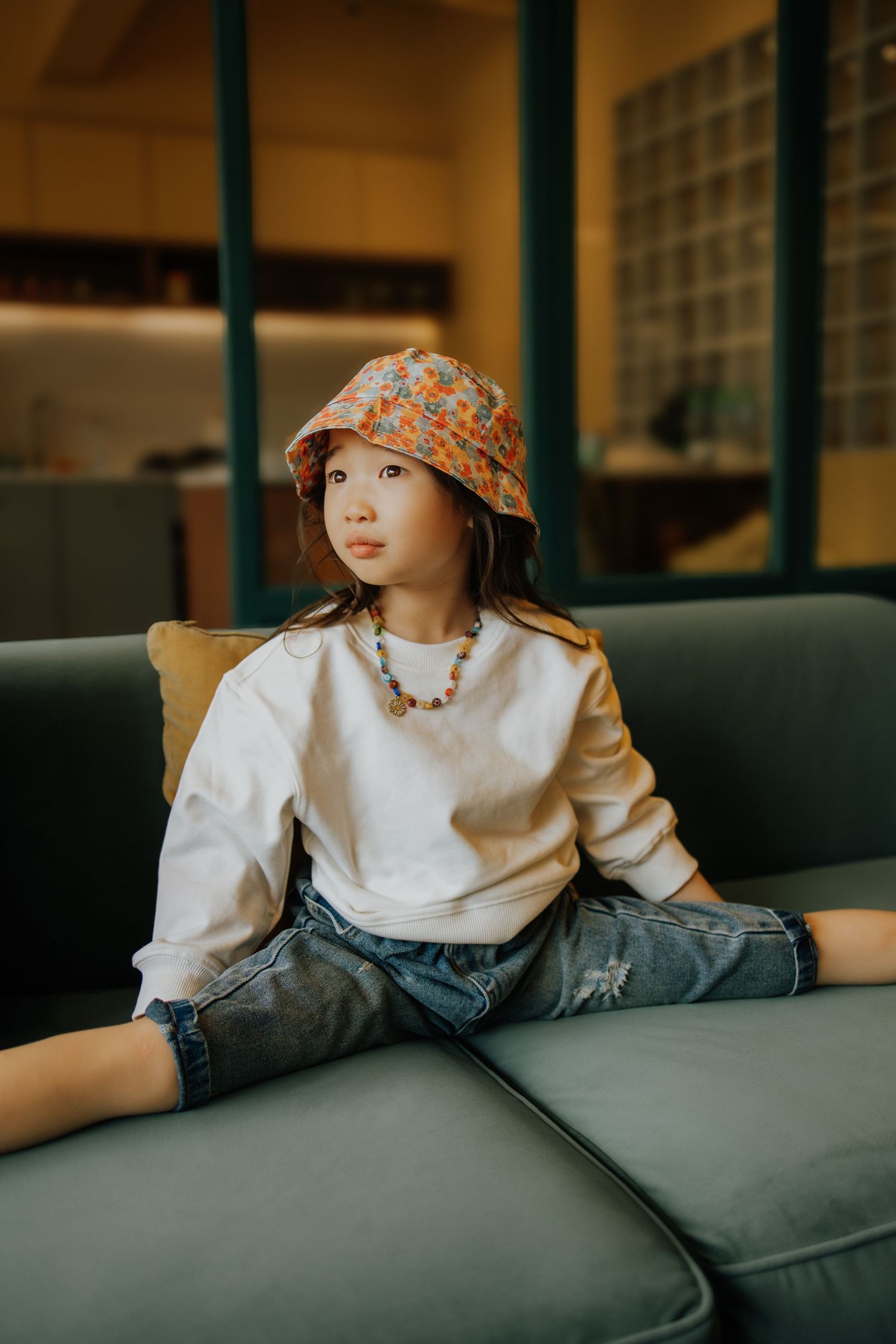 Young girl in floral bucket hat, white sweatshirt, and denim shorts sitting on a green sofa indoors.