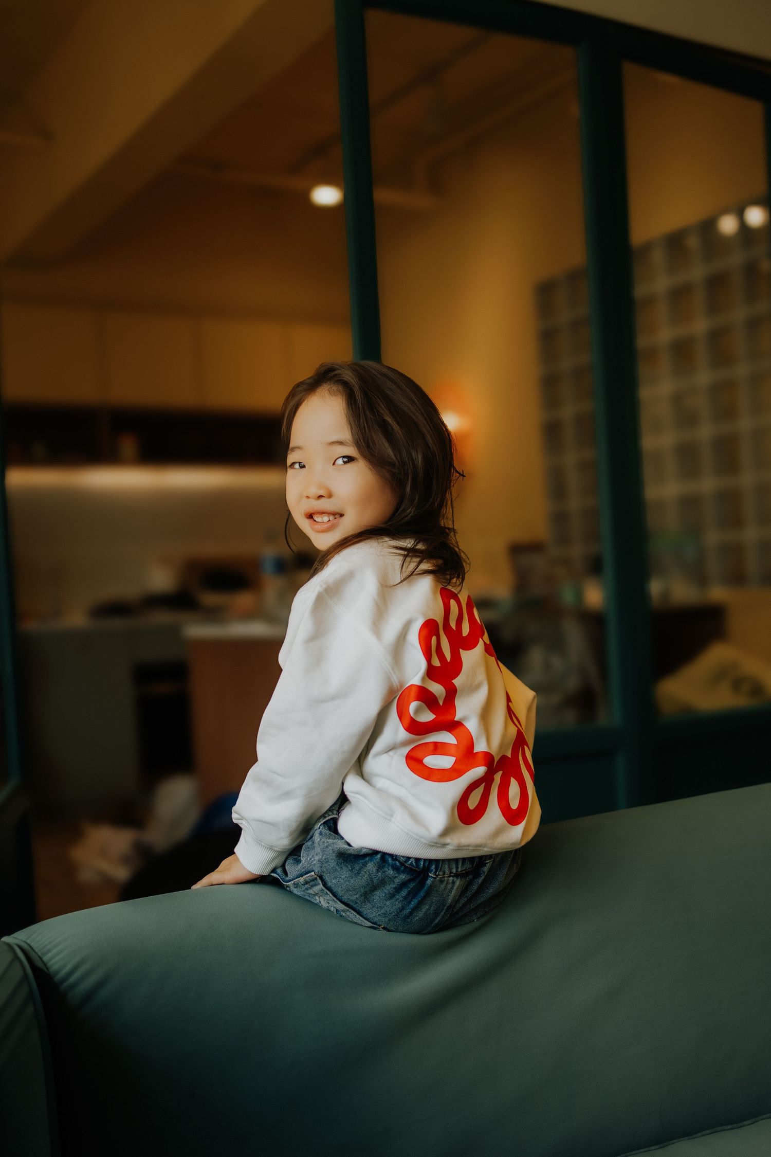A smiling young Asian girl sits on a green sofa, wearing a white sweatshirt with red design and jeans, in a warmly lit room.
