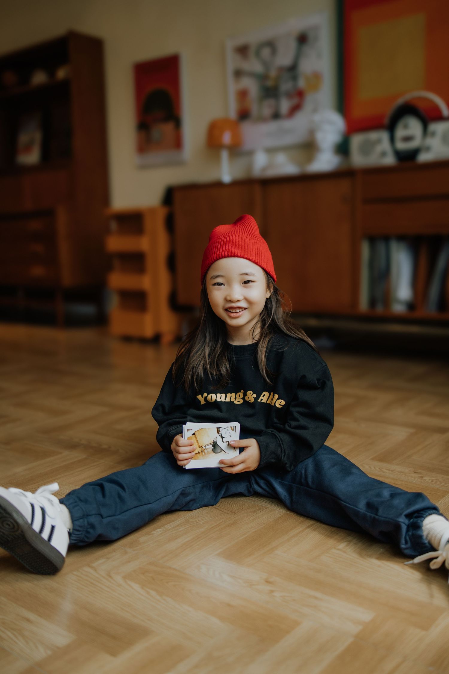 A smiling young girl in a red beanie and black sweatshirt does the splits on a wooden floor while holding a photo.