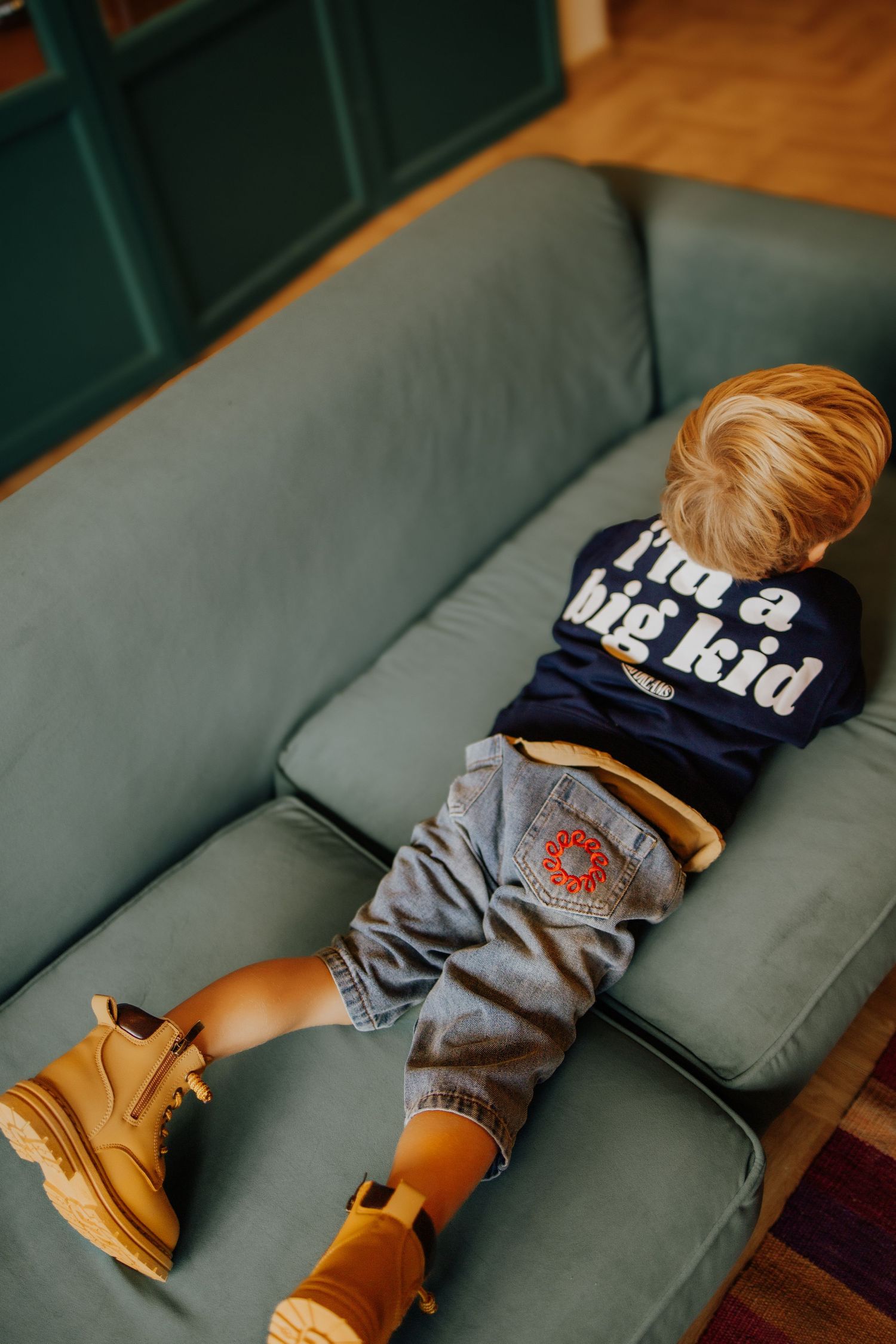 Toddler boy wearing 'I'm a big kid' shirt, jeans, and tan boots, lying on a teal sofa.