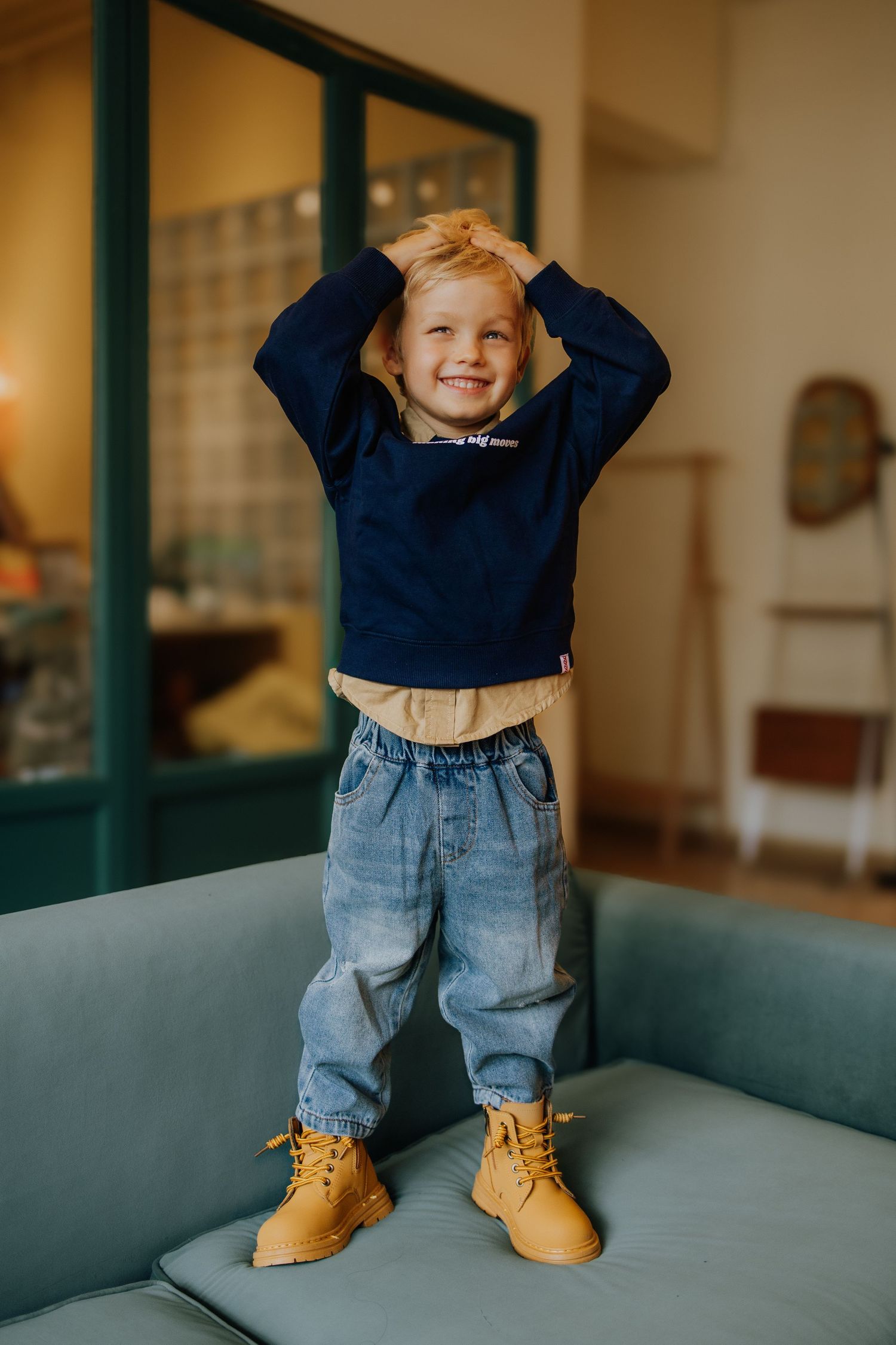 Smiling toddler boy standing on sofa, wearing navy sweatshirt, jeans, and tan boots, hands on head.