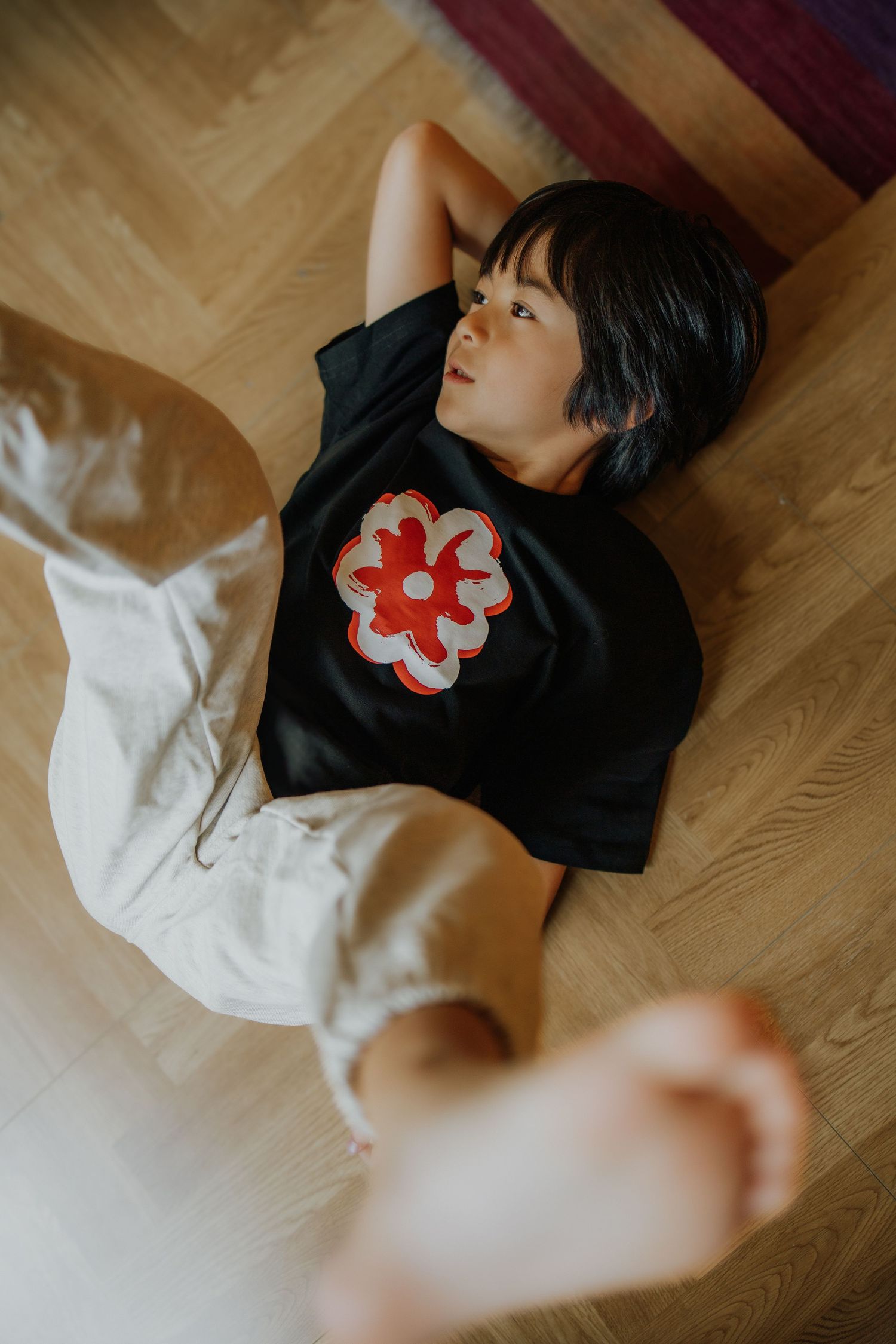 Child in black floral t-shirt and white shorts lying on herringbone wood floor, relaxed pose viewed from above.