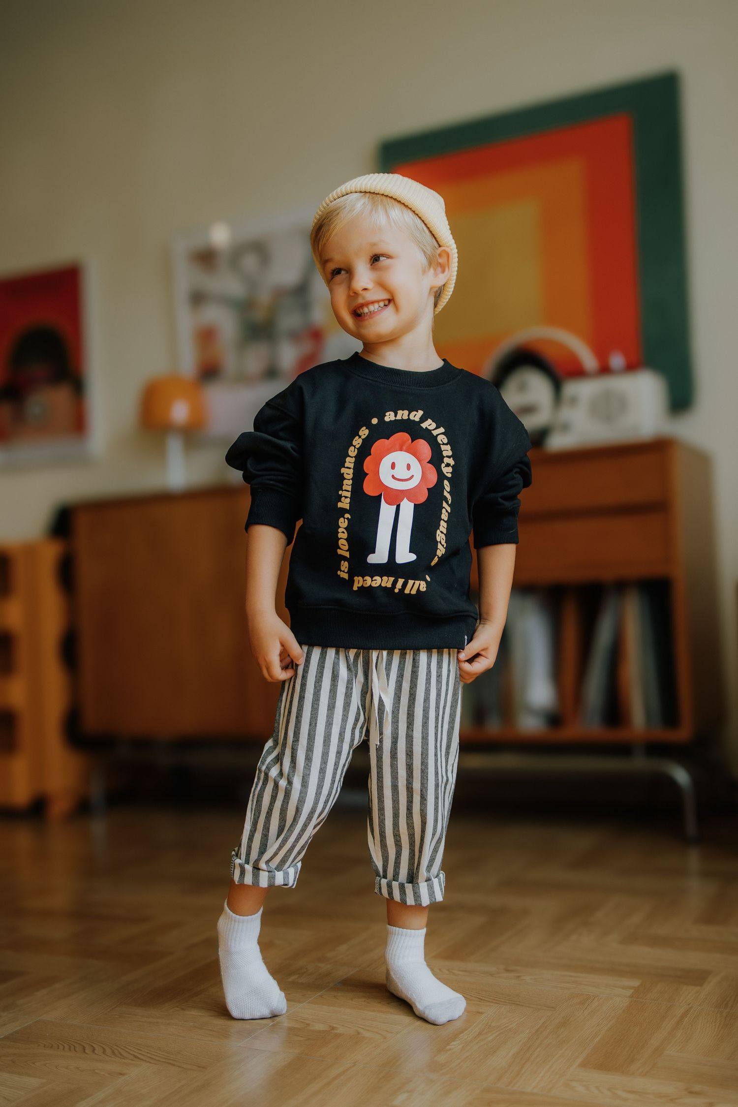 Smiling toddler boy wearing black graphic sweatshirt, striped pants, and beige hat standing indoors on hardwood floor.