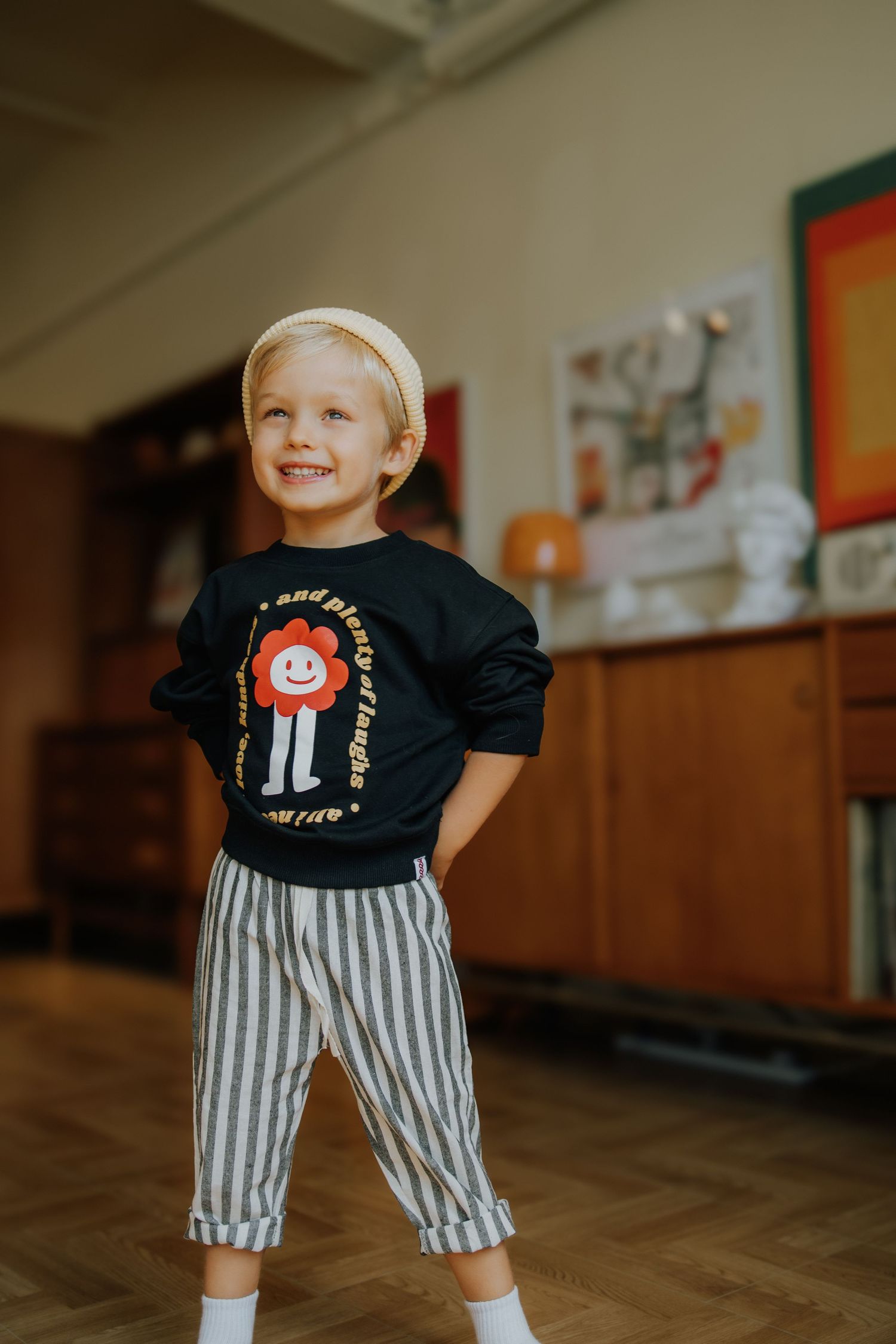 Smiling toddler in black graphic sweatshirt and striped pants with white headband posing indoors on hardwood floor.