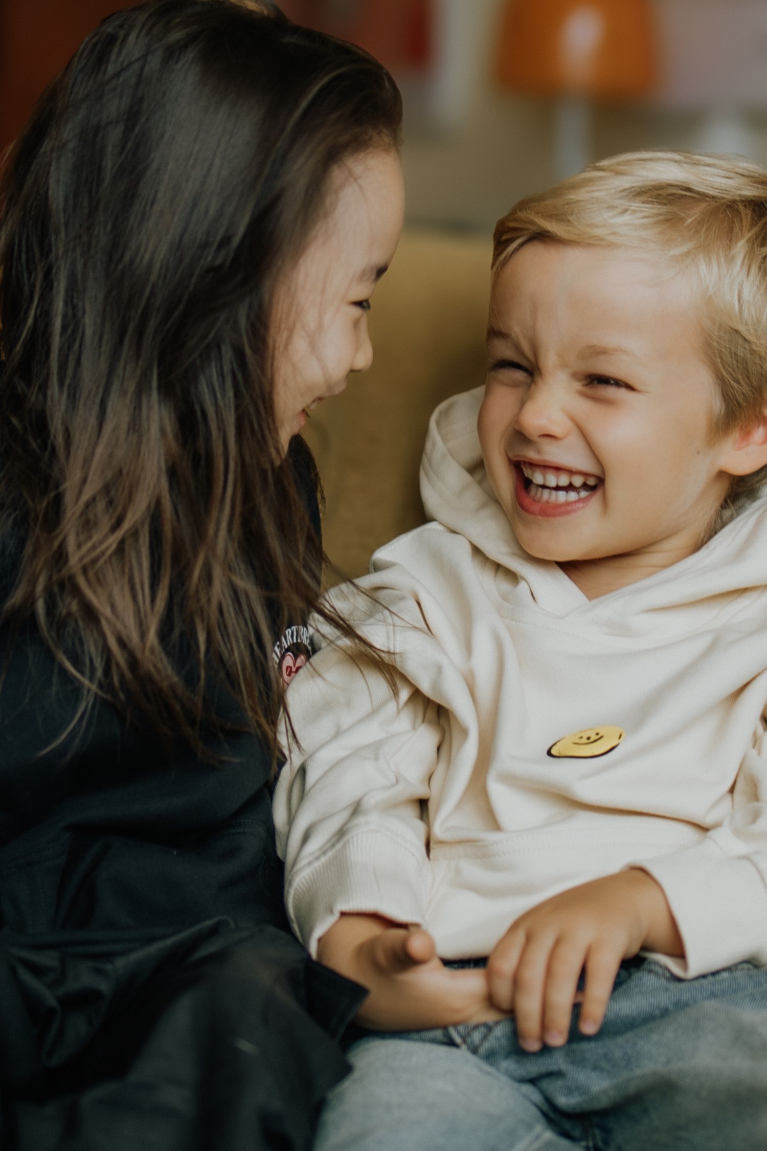 Two happy children laughing together, a girl with dark hair and a boy in a white hoodie with a smiley pin.