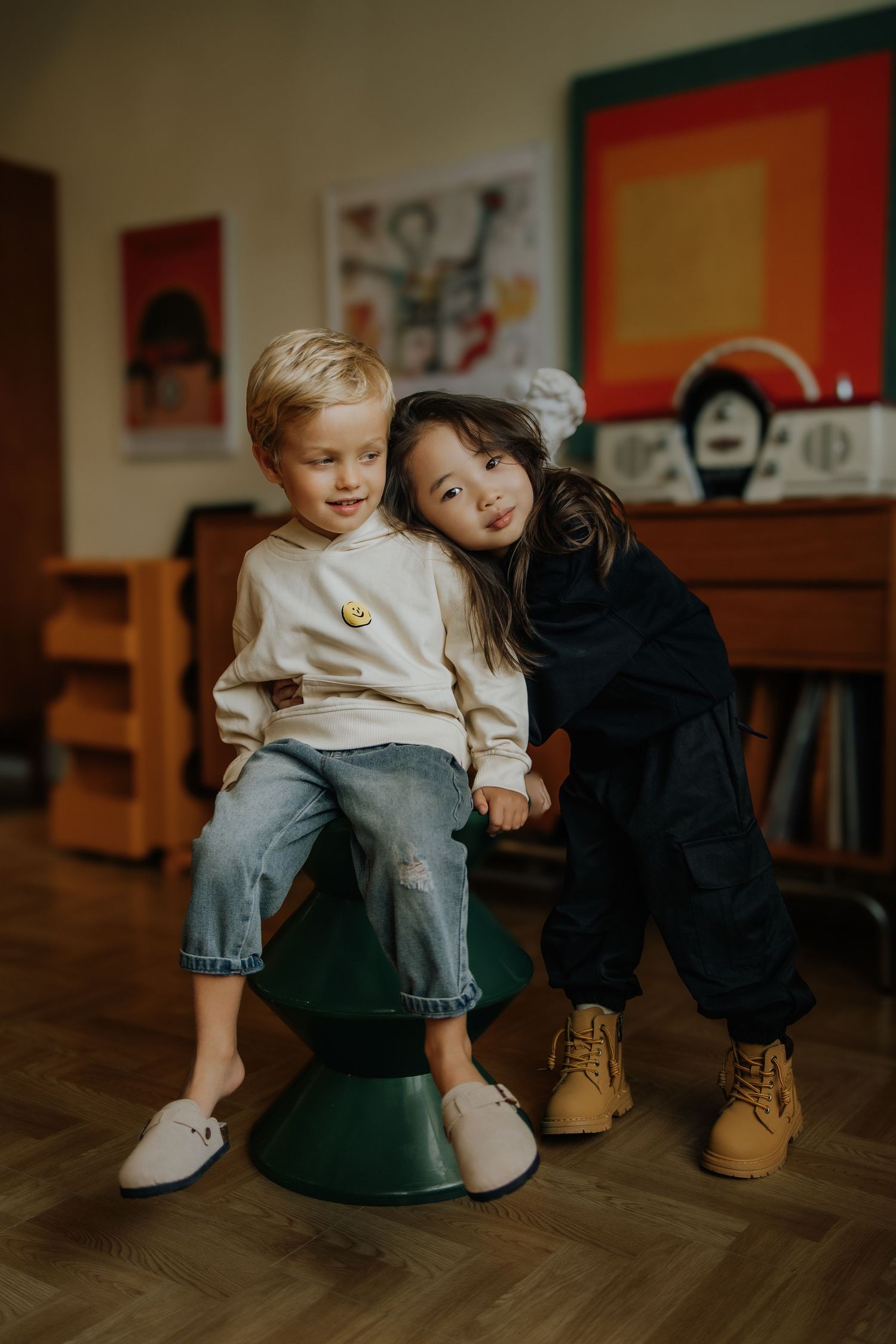 Two young children pose together in a cozy playroom, one sitting on a green stool wearing jeans and a cream sweatshirt.