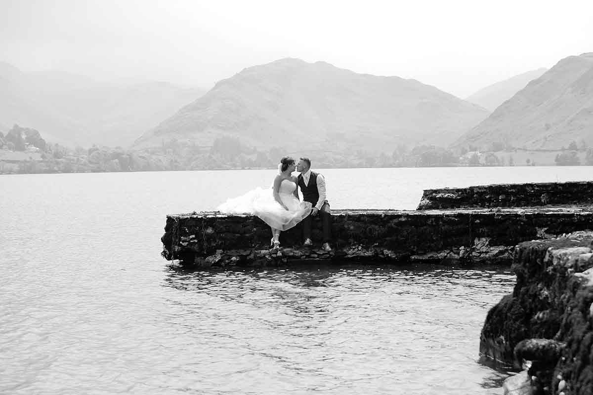 Black and white wedding photo of bride and groom kissing on rocky pier with misty mountain lake backdrop.