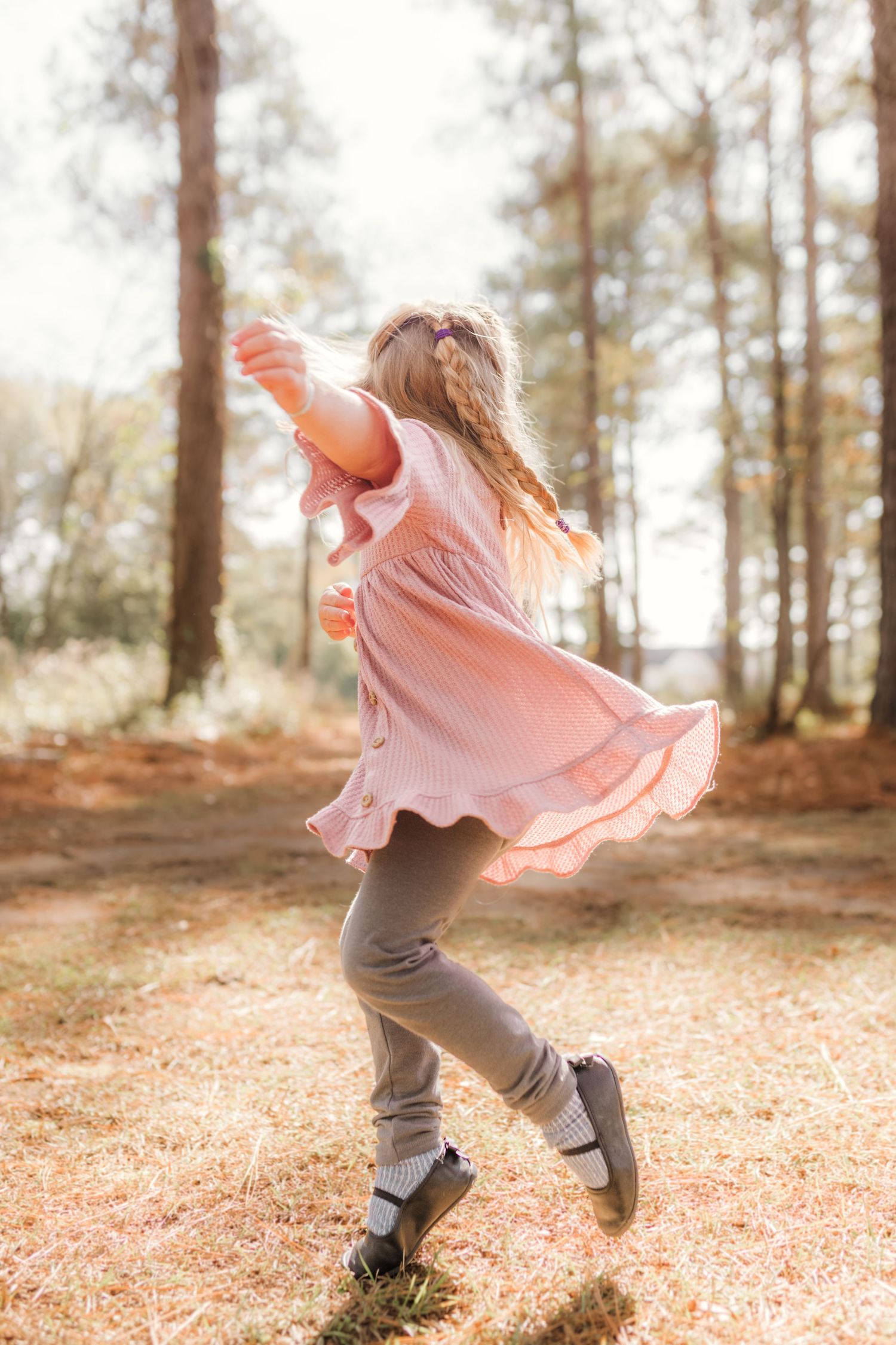 A young girl in a pink dress and gray leggings twirls joyfully in a sunlit forest clearing.