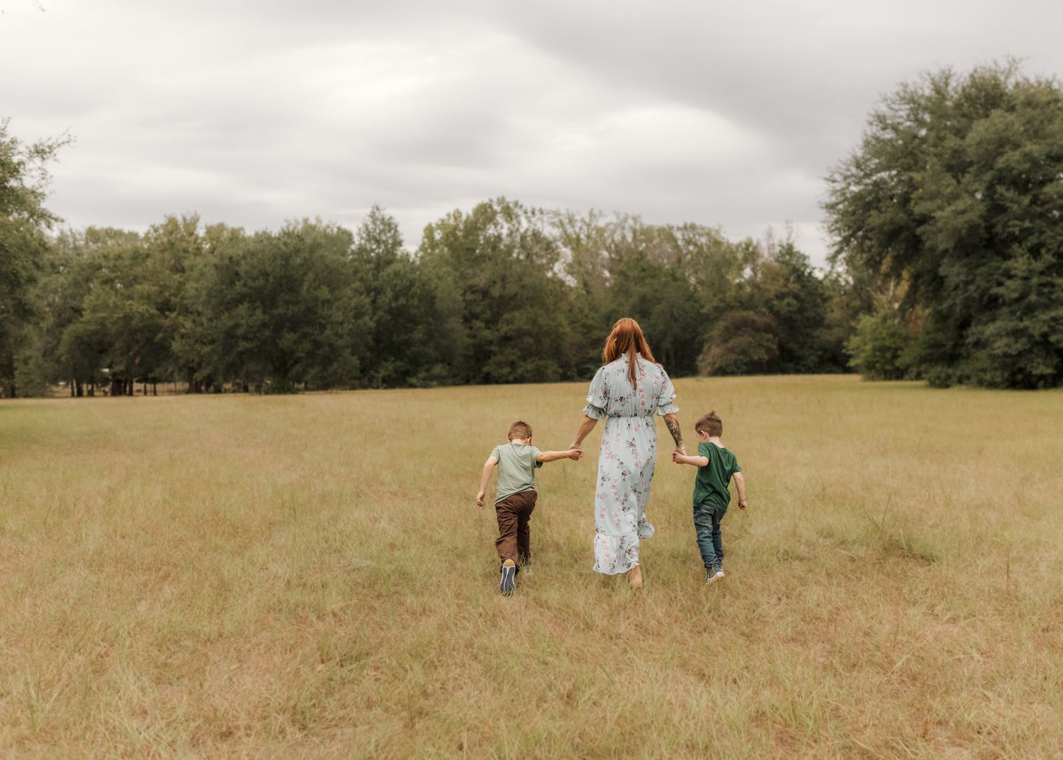 A mother walks hand-in-hand with two children across a grassy meadow surrounded by trees.