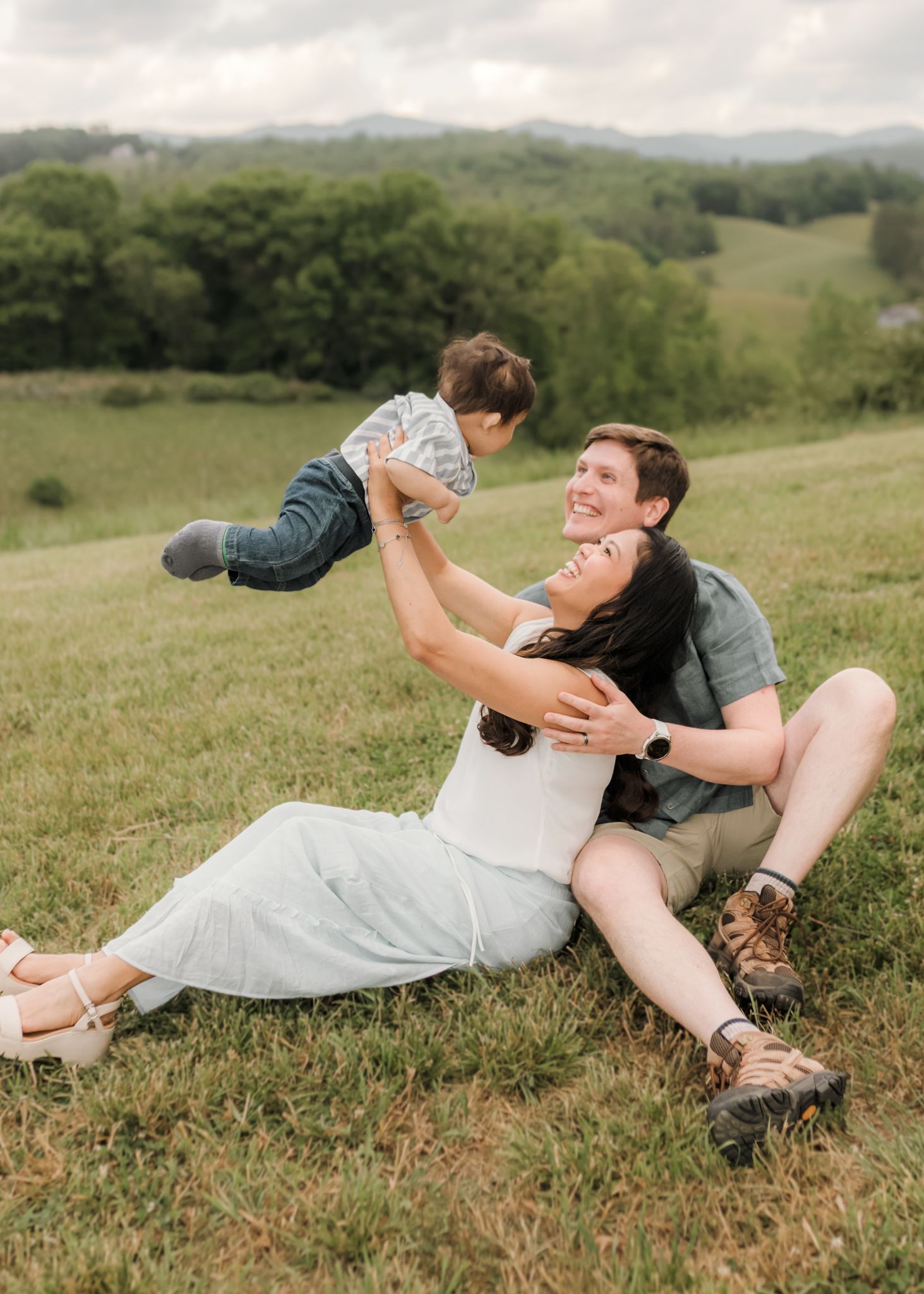 A joyful family sits on a green hillside, mom lifting toddler in air while dad smiles beside them.