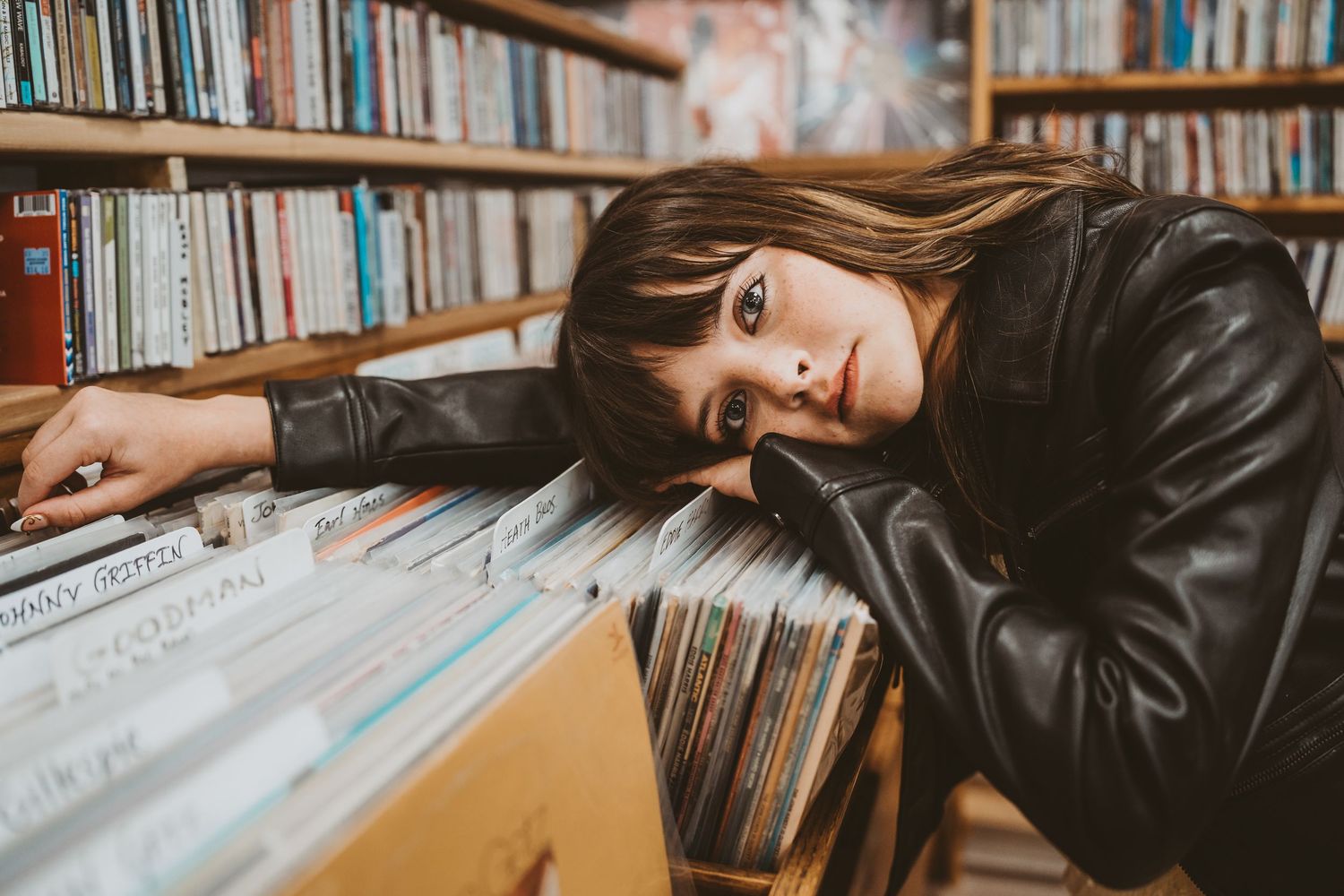 Woman in leather jacket resting head on vinyl records in a music store surrounded by bookshelves.