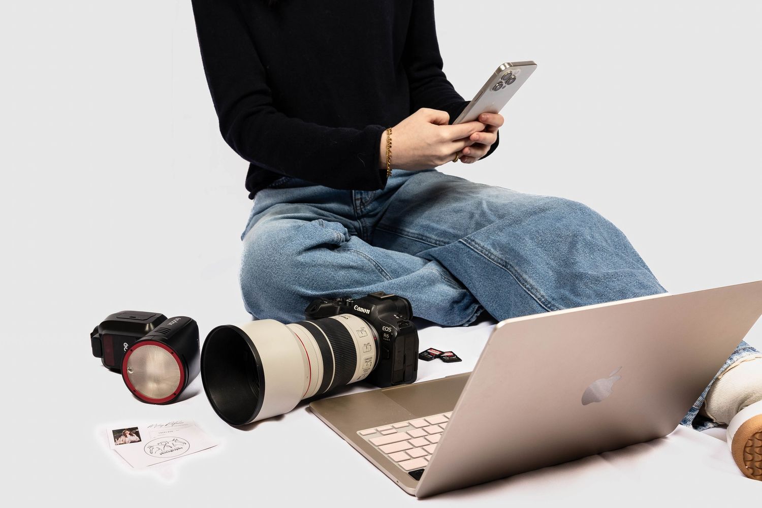 Person in black sweater and jeans sitting cross-legged using smartphone surrounded by camera gear and laptop.