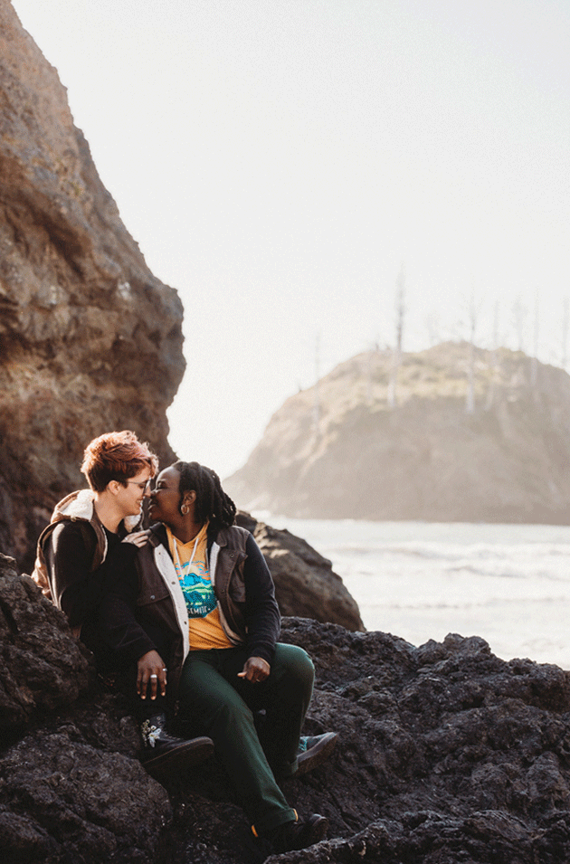 A romantic couple shares an intimate moment sitting on coastal rocks with ocean waves and misty cliffs in the background.