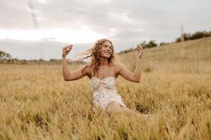 A joyful woman in a floral dress sits in a golden wheat field, arms raised, smiling under a cloudy sky.