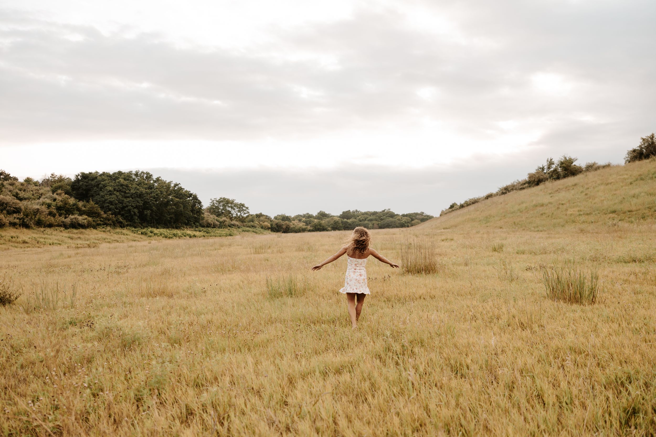 A young girl in a white dress walks through a golden wheat field under a cloudy sky.