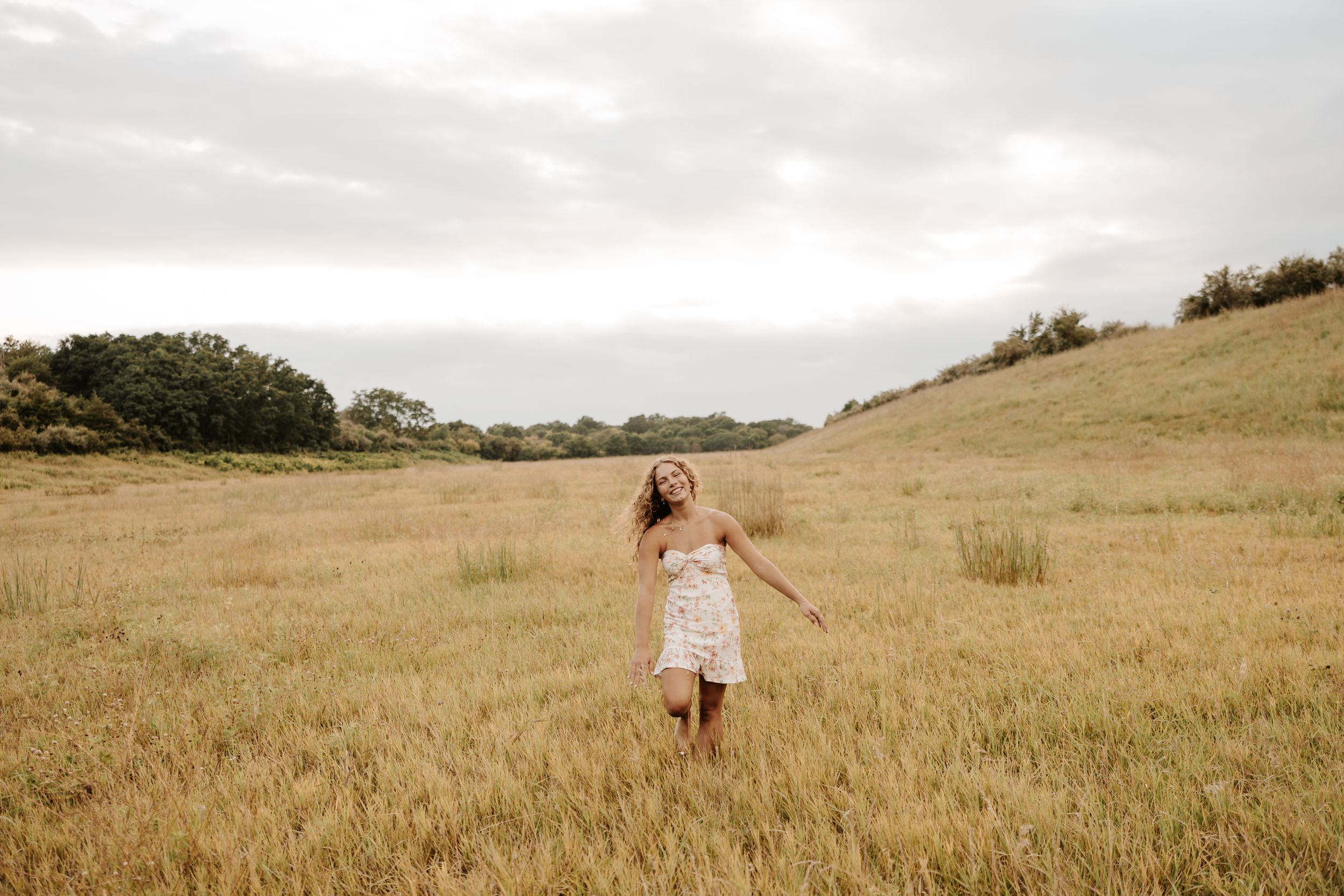 A young woman in a white dress walks freely through a golden wheat field under a cloudy sky.