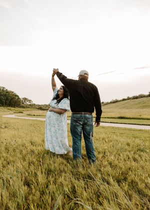 A couple dances in a lush green field, the man in black shirt and jeans twirling the woman in a floral dress.