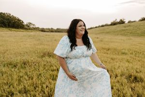 Woman in floral blue dress posing in golden field at sunset, smiling gracefully.