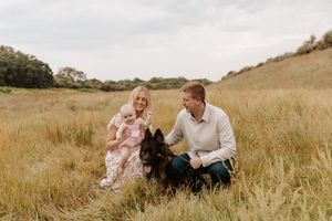 Family with young child and German Shepherd dog sitting in golden meadow grass for outdoor portrait photo.