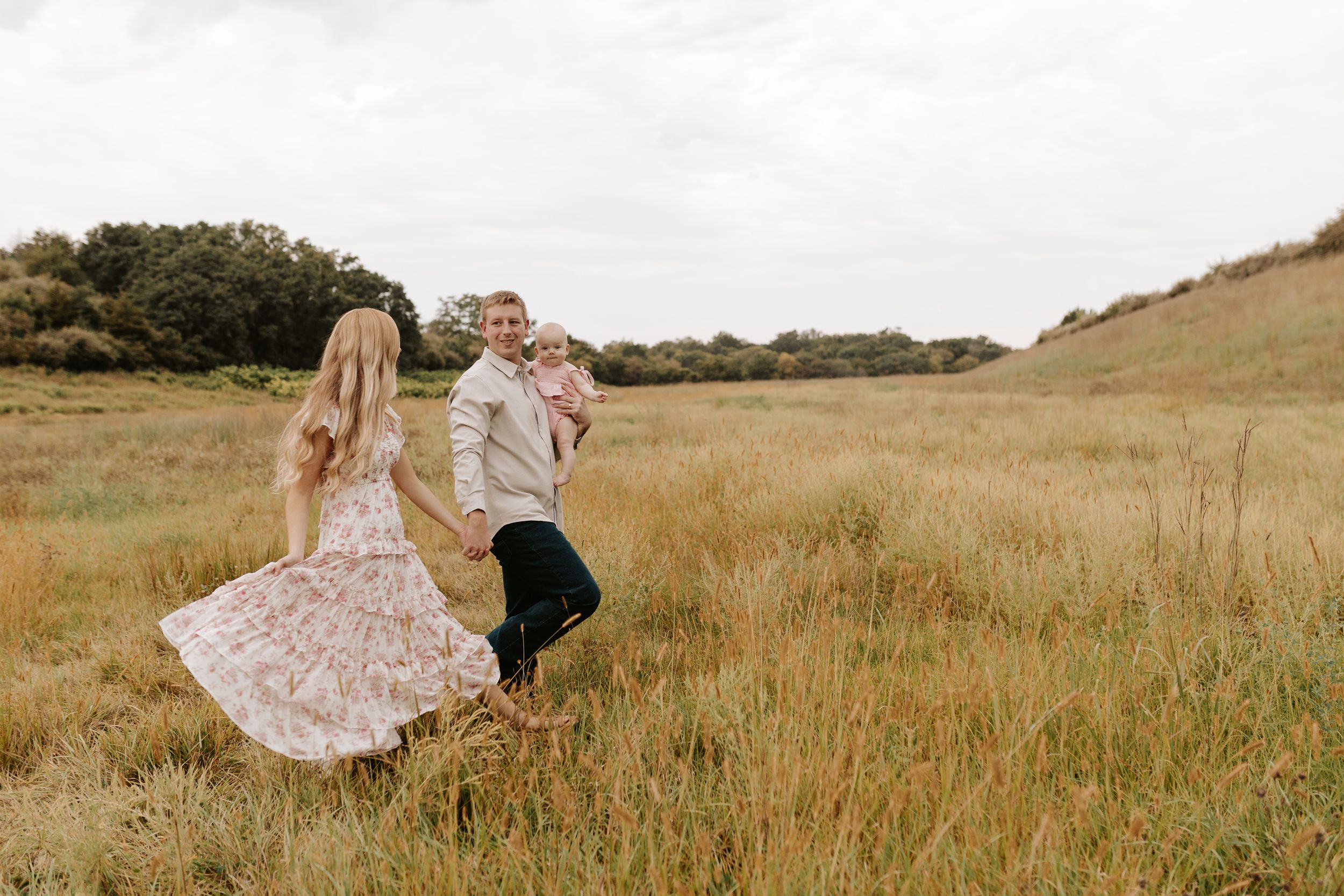 Couple dancing in a golden grass field, woman in floral dress, man in tan jacket, trees in background.
