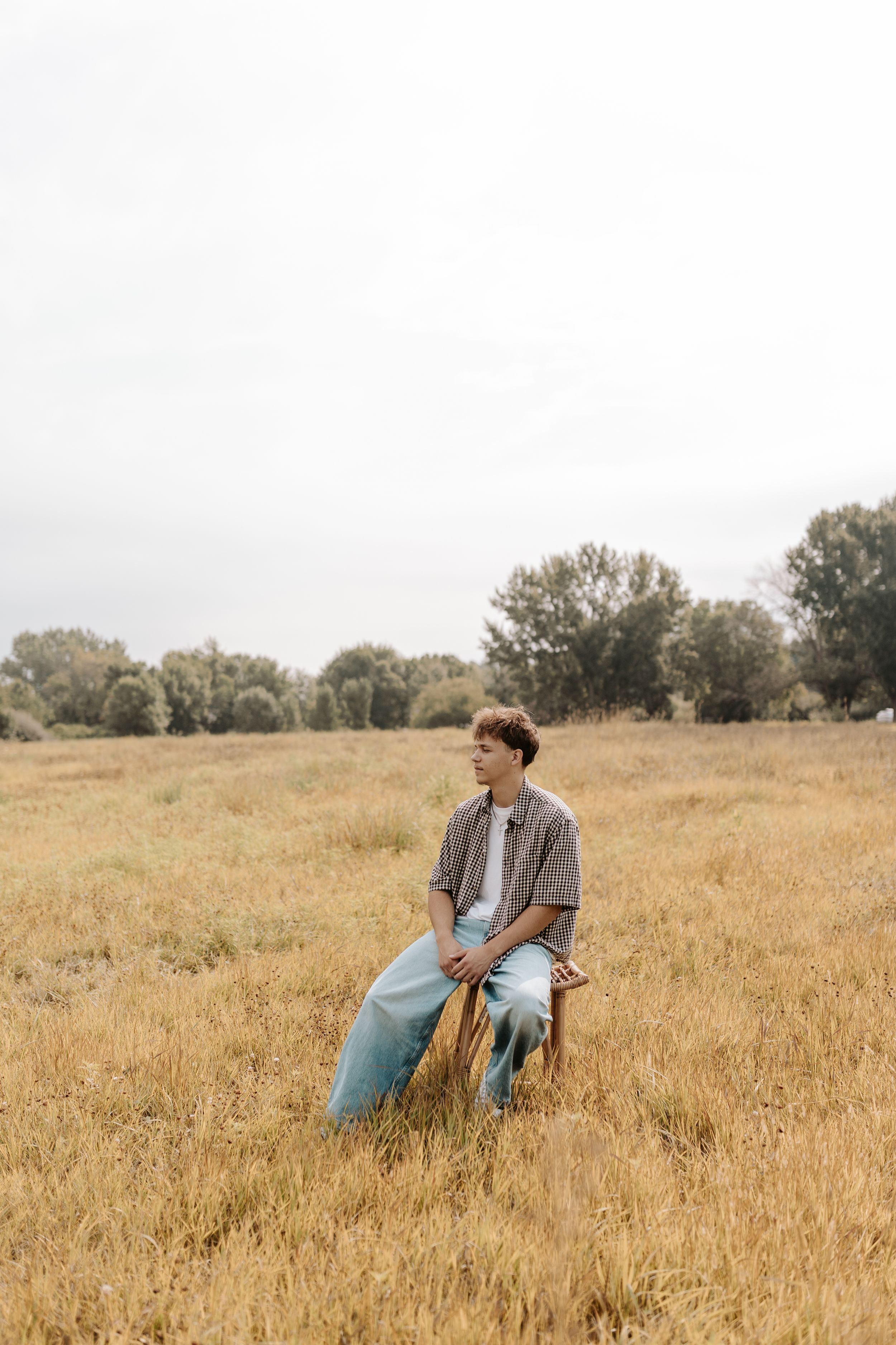 Young man sitting on a chair in a golden field, wearing a gray shirt and blue jeans, with trees in the background.