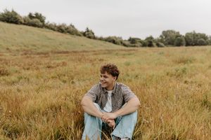 Young man smiling while sitting in a golden grass field on a cloudy day, wearing a plaid shirt and jeans.