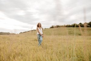 A young woman with curly hair walks through a golden meadow in jeans and white top under a cloudy sky.