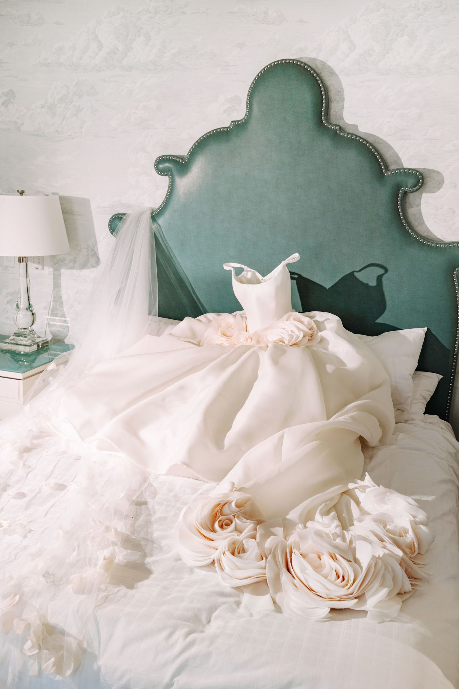 Elegant white wedding dress with large roses displayed on a bed with a teal velvet headboard in a bright bridal suite.