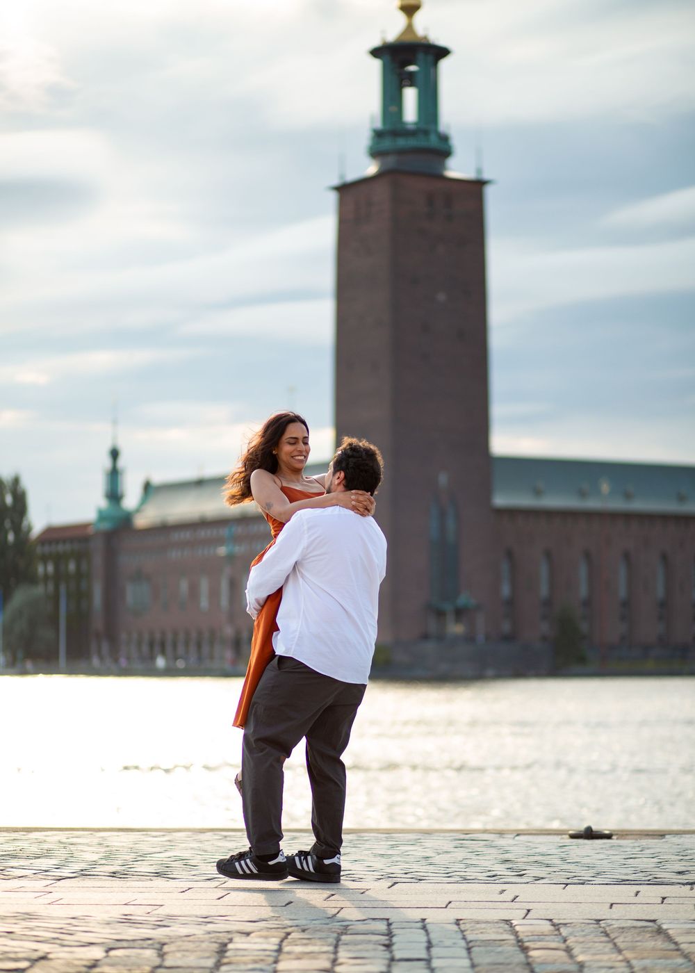 Couple embracing romantically by the waterfront with Stockholm City Hall tower in the background.