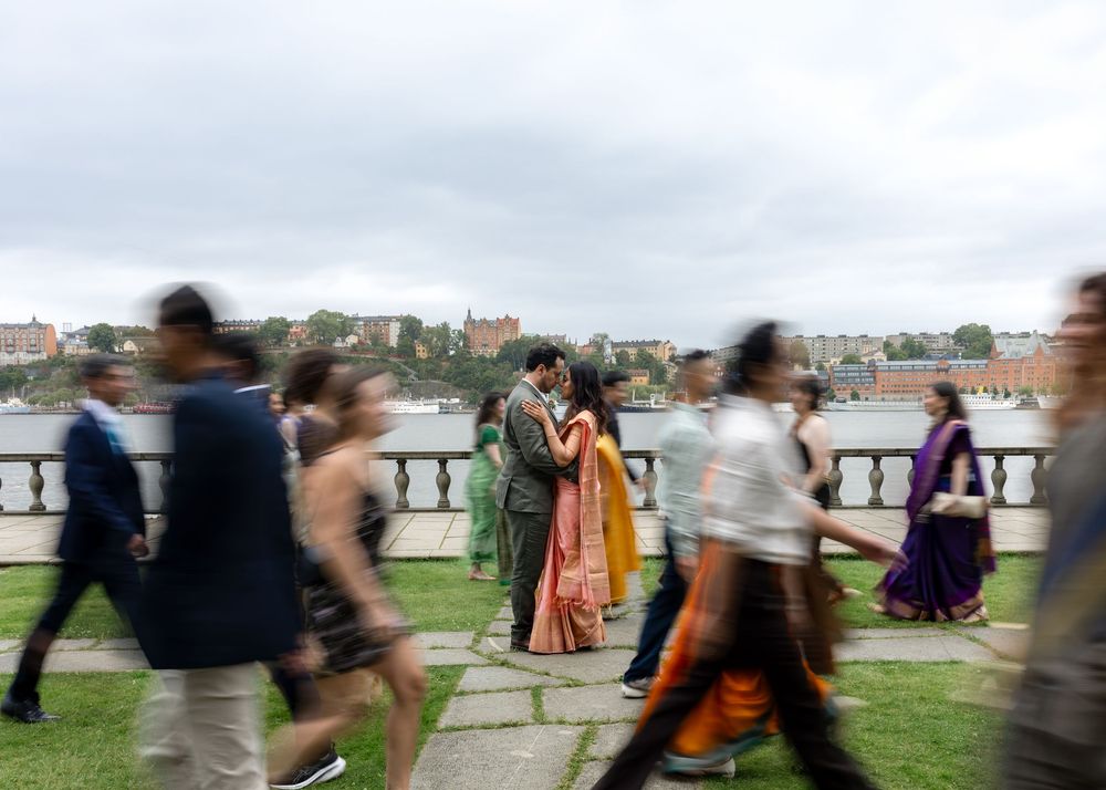 Couple in traditional Indian attire share a romantic moment amid blurred moving guests at an outdoor waterfront celebration.