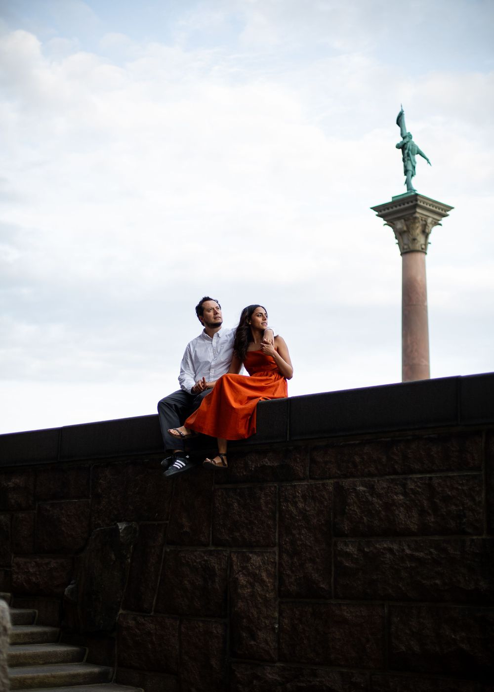 Couple sitting on a stone wall in orange dress and white shirt with a green statue column against cloudy sky.