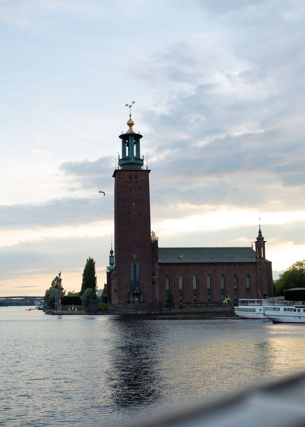 Stockholm City Hall brick tower rises beside calm waterway at dusk with boats docked nearby.